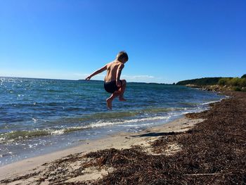 Rear view of shirtless boy jumping at beach against blue sky