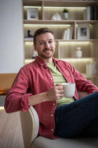Positive happy man smiling holding mug of tea sitting in chair looking camera at comfort cozy home.