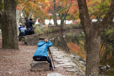 Rear view of woman sitting with tripod on rock by pond