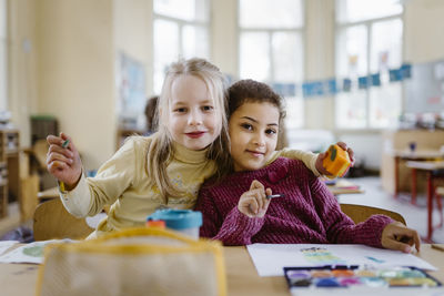 Portrait of schoolgirl with arm around female friend in classroom