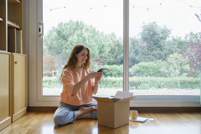 Young woman using phone while sitting on window at home