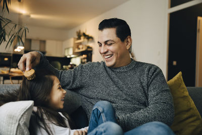 Smiling father combing daughter's hair while sitting on sofa at home