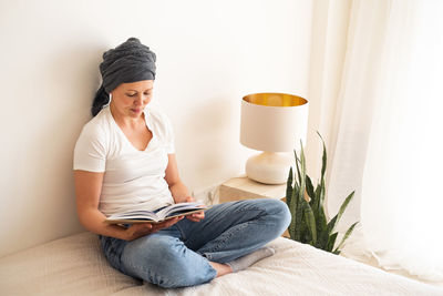 A middle-aged woman sits on a bed in her room reading a book.