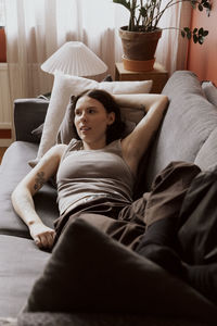 Young woman with hand raised looking away while lying down on sofa in apartment