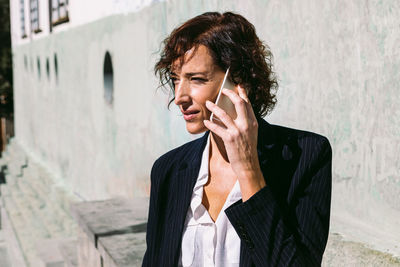 Positive adult female executive worker wearing classy outfit standing with hand on waist and talking on cellphone in sunny day