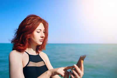 Beautiful young woman using smart phone against sea