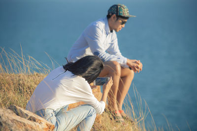 Woman photographing man sitting on mobile phone against sky