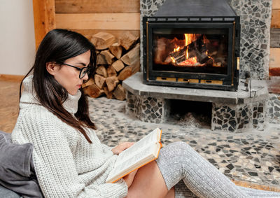 Young woman sitting by fireplace in cozy cabin, reading a book.