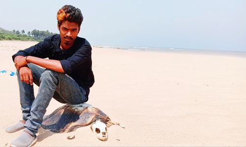 Young man sitting on dead turtle skeleton at beach against sky