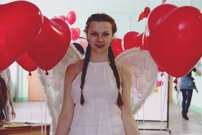 Portrait of smiling young woman wearing costume wings while standing amidst red balloons