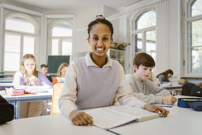 Portrait of happy schoolgirl sitting at desk with male friend in classroom