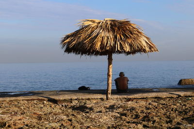 Man sitting under thatched roof at beach against sky