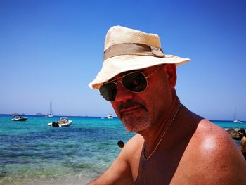 Shirtless man wearing hat and sunglasses at beach against blue sky on sunny day