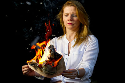 Young woman holding burning newspaper against black background