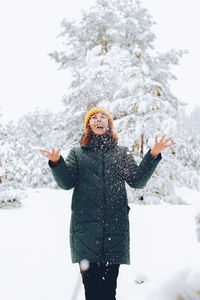 Cheerful girl with red hair in warm clothes playing with snow outdoors near the beautiful forest