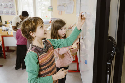 Boy writing on whiteboard while solving maths problem by girl in classroom