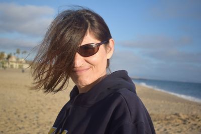 Portrait of woman with tousled hair wearing sunglasses at beach against sky