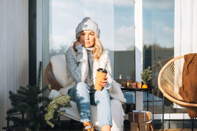 Smiling woman with blonde hair in winter clothes with coffe cup sitting near country house in winter