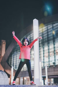 Portrait of cute girl with arms raised standing on retaining wall against built structure