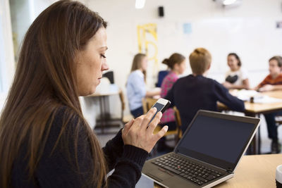 Teacher using mobile phone at classroom with students in background
