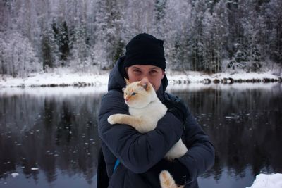 Portrait of young man carrying cat while standing against lake in forest