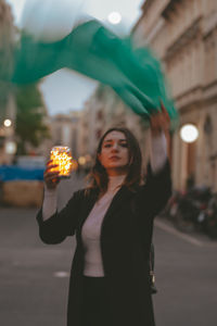 Portrait of woman with green scarf and illuminated jar standing on city street at dusk