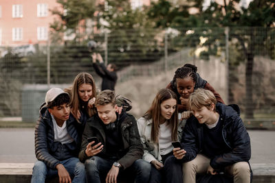 Multi-ethnic teenage friends using smart phones while sitting at park in city