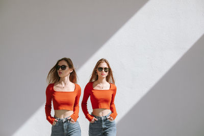 Two sensual young twin sisters in sunglasses and identical clothes posing in contrast sunlight.