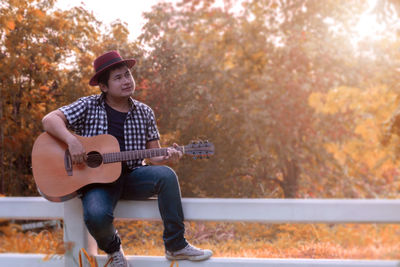 Thoughtful young man sitting and playing guitar on fence