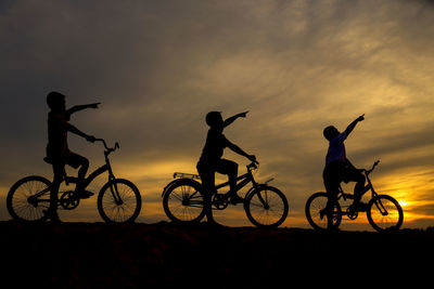 Silhouette boys pointing while riding bicycle against orange sky