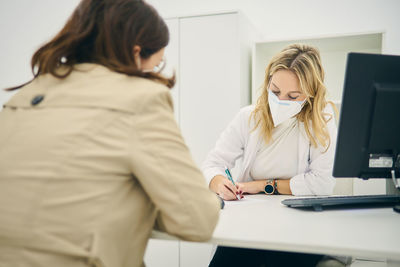 Female dermatologist with face mask sitting at table with client and taking notes on paper while writing prescription for buying cosmetic products during coronavirus pandemic