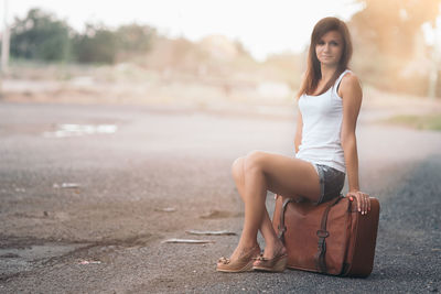 Portrait of young woman sitting over suitcase on road