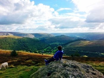 Side view of boy  sitting on rock overlooking landscape against cloudy sky