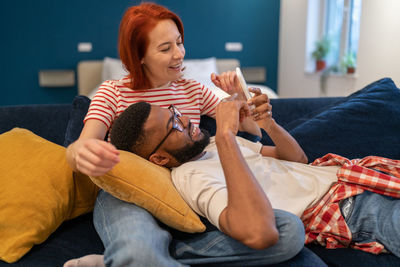 Multi ethnic happy couple man and woman look at phone lying on couch in living room.