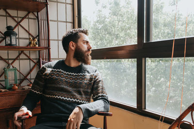 Young man looking away while sitting on window