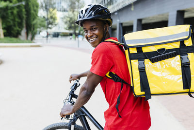 Happy young delivery man wearing backpack sitting on bicycle