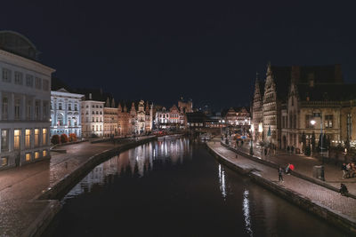 Illuminated street by buildings against sky at night