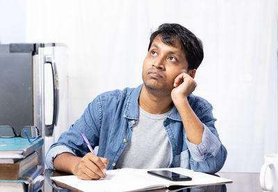 An indian young male looking upwards and thinking while studying at home on white background