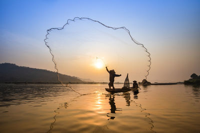 Silhouette people fishing while standing in boat on lake against sky during sunset