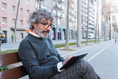 Handsome mature man in eyeglasses using digital tablet on bench in the street