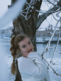 Portrait of young woman standing in snow by tree outdoors