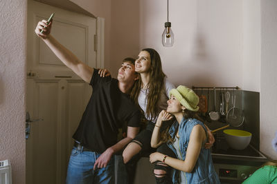 Young man taking selfie with smiling female friends in kitchen at home