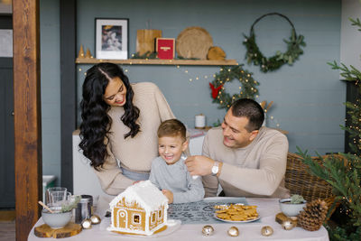 The family bakes a gingerbread house for christmas with sweets, the father puts icing on son's nose