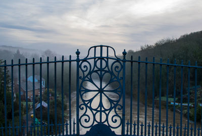 Closed gate in front of river amidst trees against cloudy sky