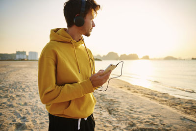 Young man using listening music at beach against sky during sunset