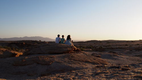 Rear view of friends sitting on rock formation against clear sky