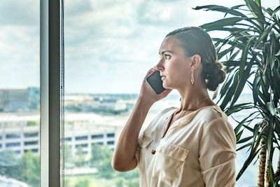 Woman talking on mobile phone against glass window in office