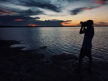 Silhouette woman photographing sea against sky during sunset