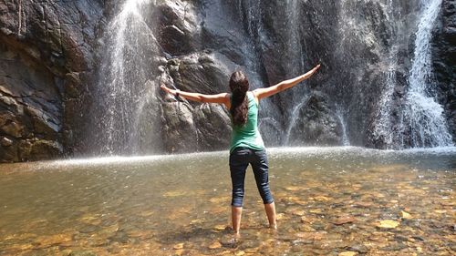 Full length rear view of woman with arms outstretched standing against waterfall