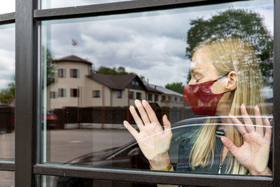 Depresive woman in face mask looking out the window, staying home in quarantine, covid-19 lockdown